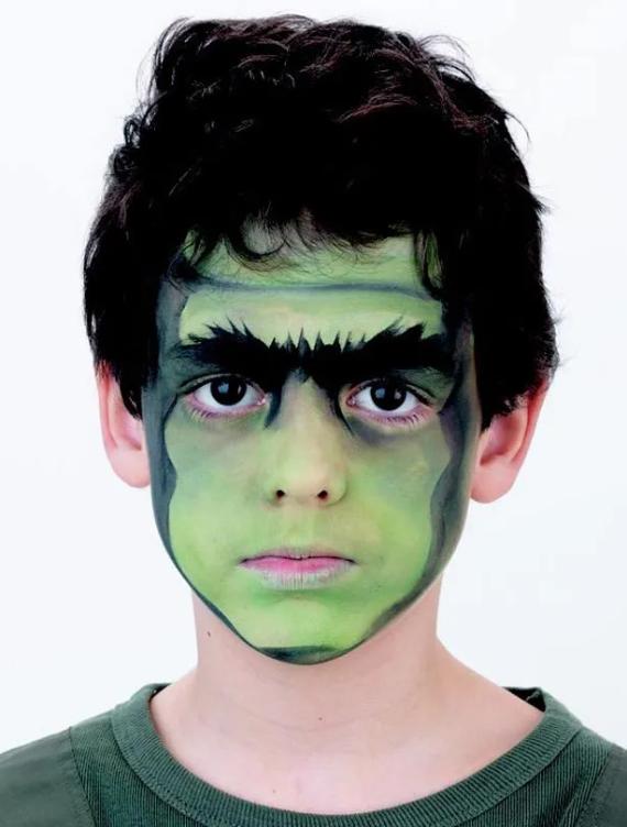 Young boy with simple scary Halloween makeup for kids feathering a face painted green as the Hulk, complete with dark bushy eyebrows and intense expression. Wears a green shirt, conveying a playful, dramatic tone.