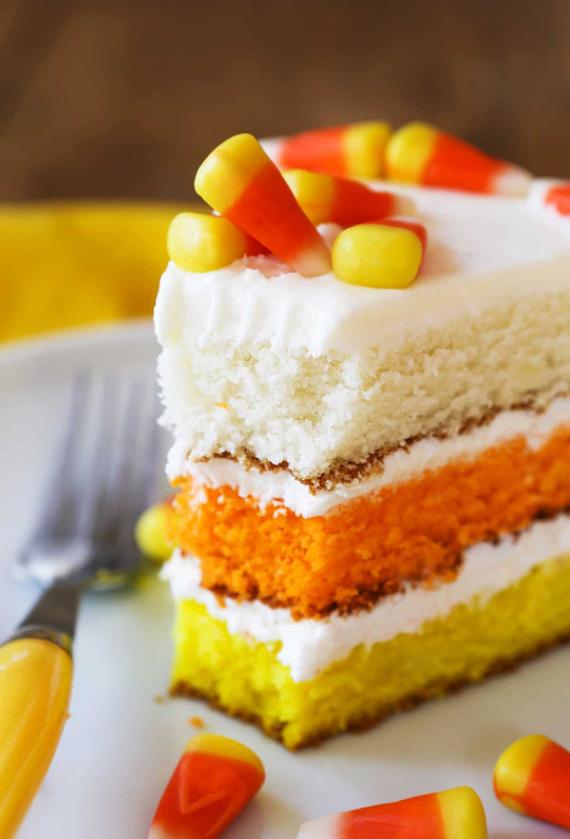  A slice of Halloween layer cake featuring white, orange, and yellow cake layers with white frosting, topped with scattered candy corn pieces on a white plate.