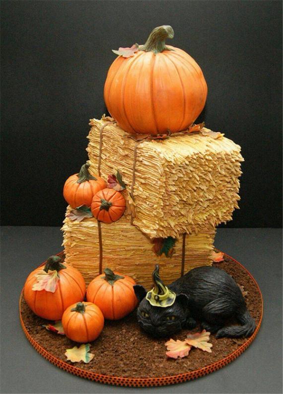 A realistic harvest Halloween cake shaped like stacked hay bales decorated with sculpted orange pumpkins, fall leaves, and a black cat wearing a witch hat.