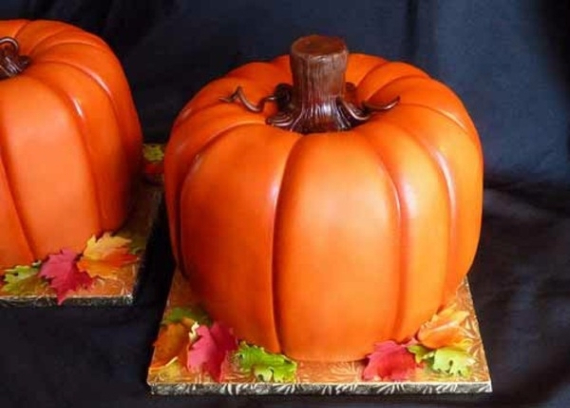 A realistic 3D sculpted pumpkin cake covered in smooth orange fondant with a textured brown stem and colorful autumn leaves at the base.