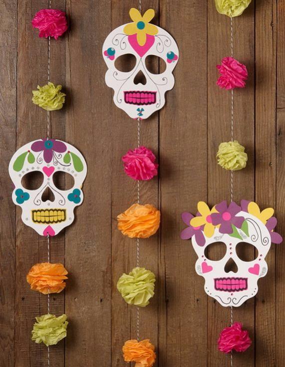 Three decorated white paper sugar skulls hanging vertically on strings alongside colorful pink, orange, and green tissue paper pom-poms against a rustic wooden wall.