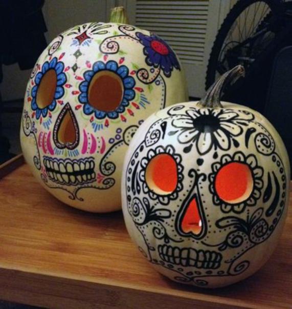  Two white pumpkins decorated as Day of the Dead sugar skulls with hand-painted colorful patterns and carved, glowing eyes resting on a wooden tray.