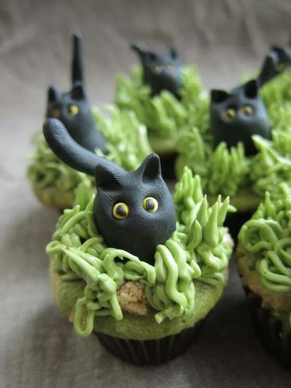 A group of cupcakes decorated with spiky green frosting grass and molded black fondant cats with yellow eyes peeking out.