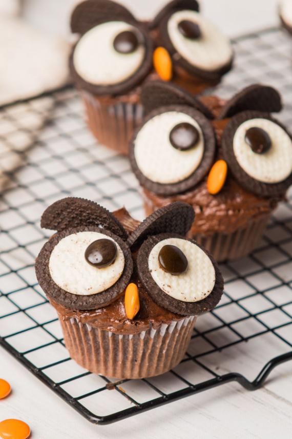  Three chocolate cupcakes decorated with Oreo cookie eyes, chocolate candy pupils, and orange candy beaks to look like owls on a wire rack.