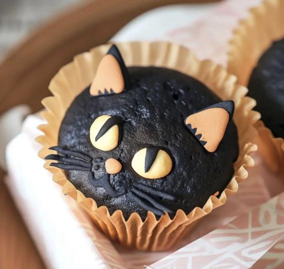  A dark black chocolate cupcake decorated like a cat face with yellow eyes, whiskers, and pointed ears in a brown wrapper.