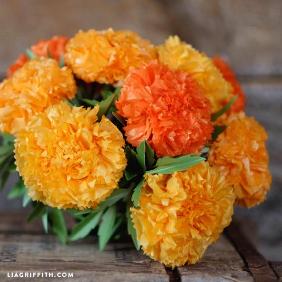 Handcrafted yellow and orange tissue paper marigold flowers with green paper leaves arranged in a bouquet on a rustic wooden surface.