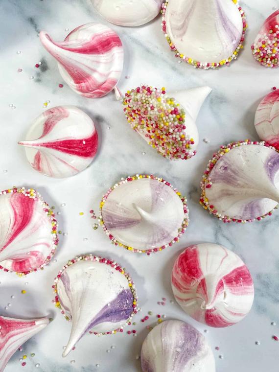 Pink and purple swirled meringue cookies shaped like witch hats, decorated with multi-colored sprinkles along the bottom rims, resting on a white marble background.