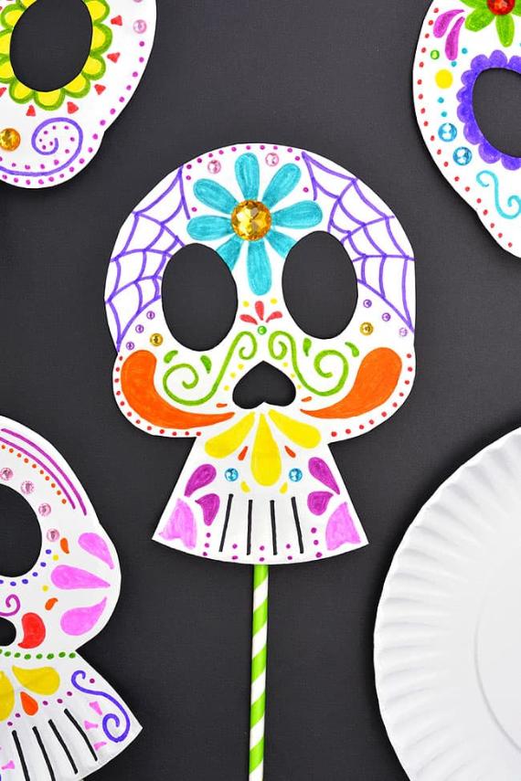 Handcrafted Day of the Dead sugar skull masks made from paper plates, decorated with colorful markers, gems, and attached to green striped straws on a black background.