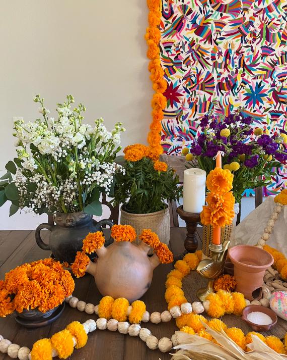 Traditional Mexican Day of the Dead altar setup with orange marigold garlands, clay pottery, and an Otomí embroidered textile backdrop.