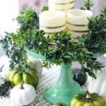 a-modern-Thanksgivving-centerpiece-of-a-green-stand-with-greenery-striped-candles-white-and-green-pumpkins-and-a-printed-runner