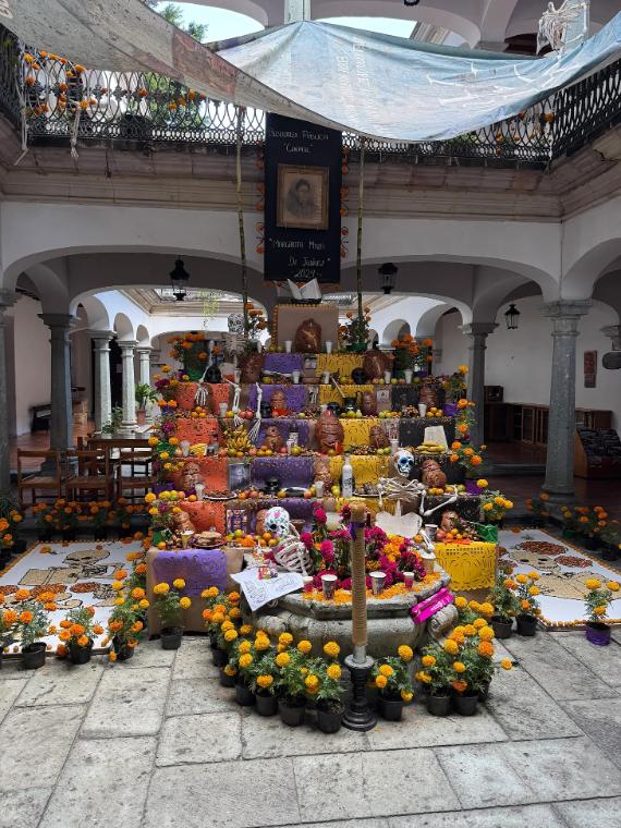 A large, multilevel traditional Mexican Day of the Dead ofrenda altar built inside a stone courtyard, decorated with orange cempasúchil marigold flowers, purple paper decorations, calavera skeleton figurines, sugar skulls, candles, and food offerings.