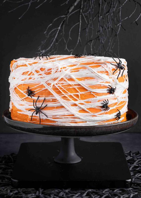 Bright orange Halloween cake covered in stretchy melted marshmallow spiderwebs and decorated with small plastic black spiders, sitting on a dark pedestal stand.