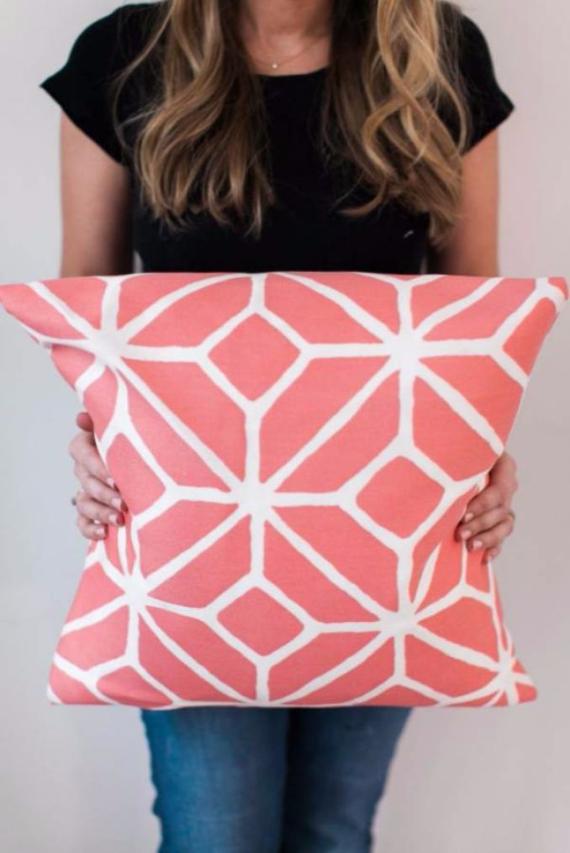 Woman in a black shirt and jeans holding a coral pillow with a white geometric pattern. Her face is not visible, with a casual and relaxed tone.