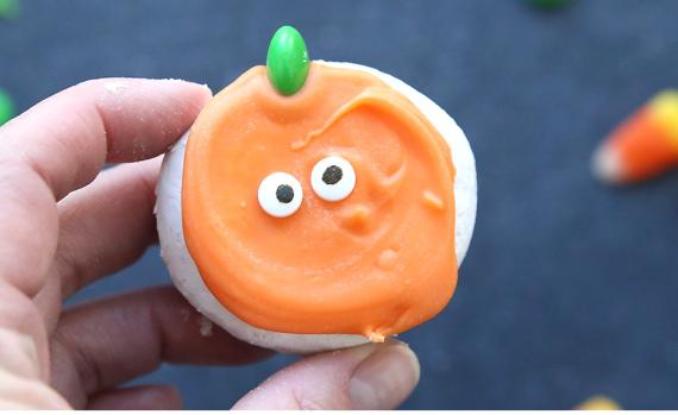 A hand holding a pumpkin-shaped cookie with eyes, showcasing a festive Halloween treat.