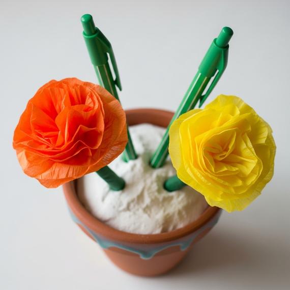 Three pens with vibrant orange and yellow paper flowers are arranged in a clay pot filled with white sand, creating a Pen Holder.