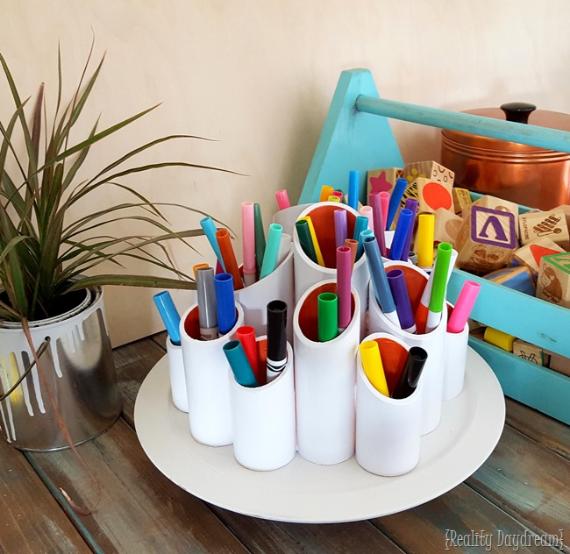 Colorful markers in a circular white pen holder on a wooden table, next to a potted plant and a blue tray with blocks, creating a lively, organized scene.