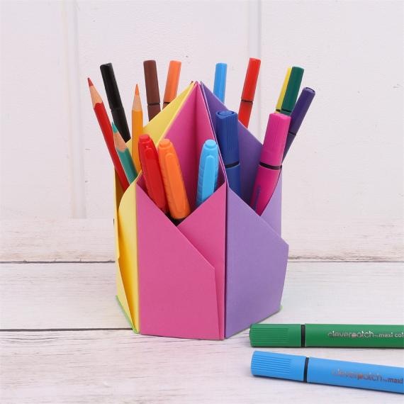 Colorful markers and pencils are neatly arranged in a colorful, Origami Pencil Holder, set on a light wooden surface, creating a cheerful and organized scene.