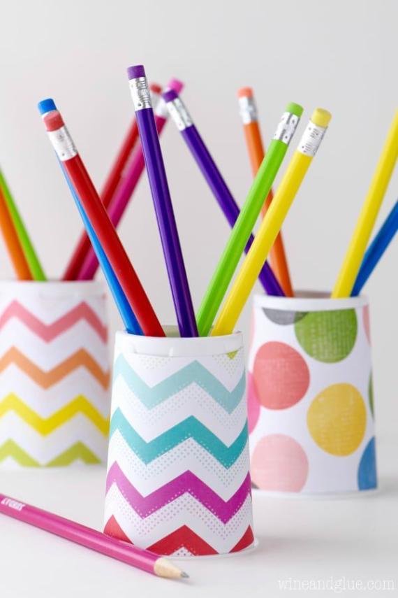 A variety of colorful pencils stand in white holders with zigzag and polka dot patterns on a white surface. The pencils' vibrant colors add a lively tone.