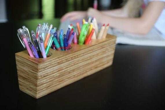 Wooden pen holder with colorful pens and markers arranged neatly on a dark table. A person, out of focus, sits writing in the background.