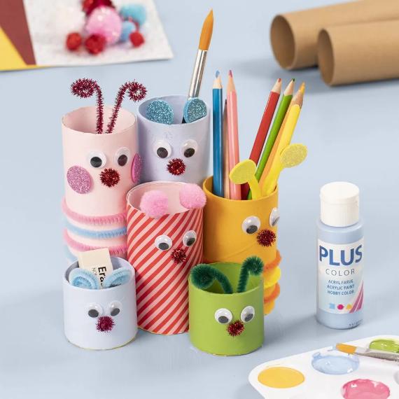 Colorful DIY pencil holders made from decorated toilet paper rolls with googly eyes and pom-poms. Paint and craft supplies are nearby on a light blue surface. Fun and whimsical tone.