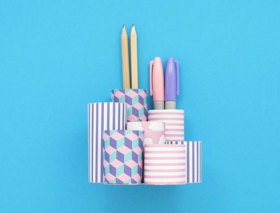 Colorful geometric pen holder on blue background. Contains two pencils, two pens, and various patterned segments in stripes and cubes. Bright, organized.