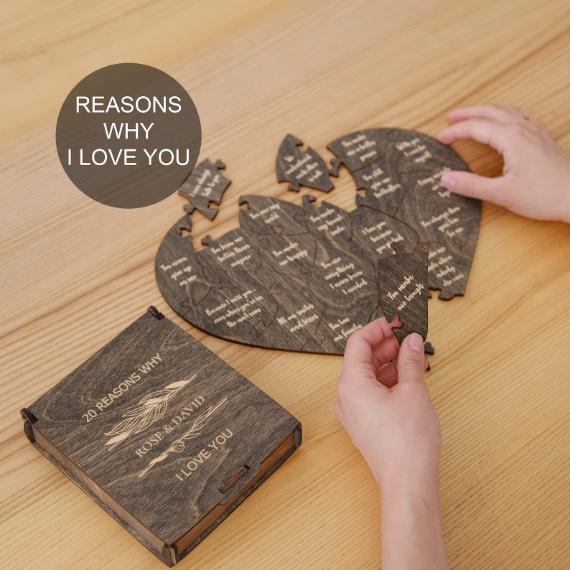 Hands assemble a heart-shaped puzzle with engraved text pieces, beside a wooden box labeled "20 Reasons Why I Love You" on a wooden table.