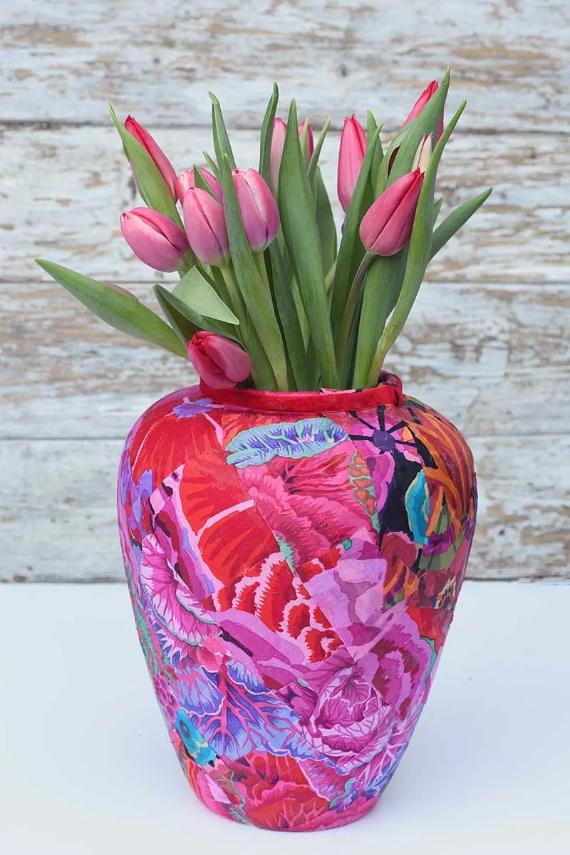 Vibrant pink tulips in a colorful, abstract-patterned vase with reds and blues, set against a rustic white wooden background, convey a lively and artistic tone.
