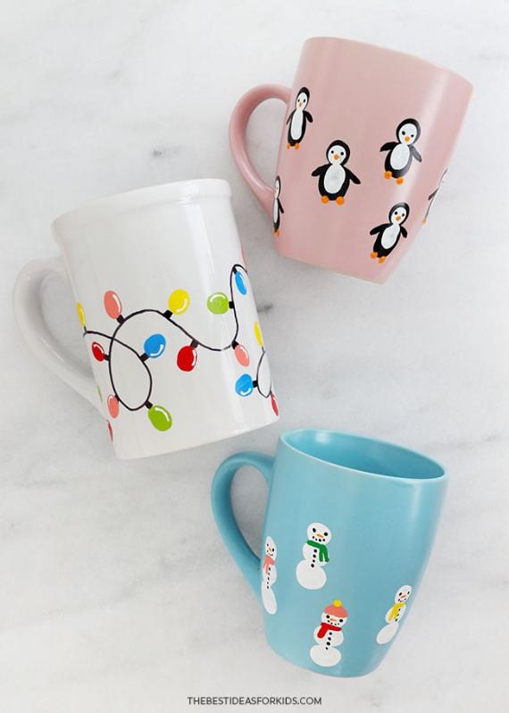 Three mugs on a white surface: one white with colorful Christmas lights, a pink mug with penguins, and a blue mug with snowmen. Festive and cheerful.