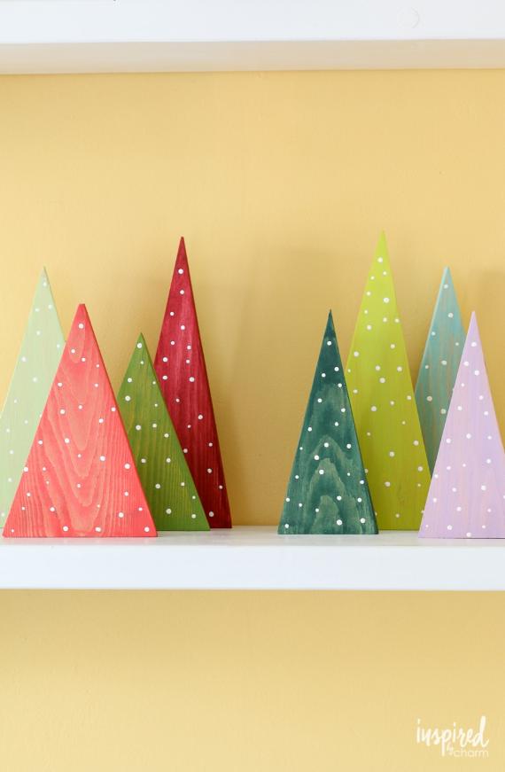 Colorful, triangular wooden trees of varying sizes with white polka dots are displayed on a white shelf against a yellow wall, creating a whimsical, festive atmosphere.