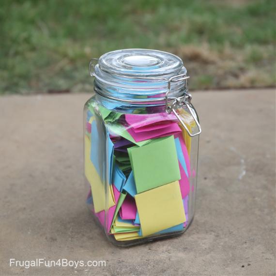 A glass jar filled with colorful folded paper notes, including pink, blue, yellow, and green, sits on a concrete surface. Grass is visible in the background.