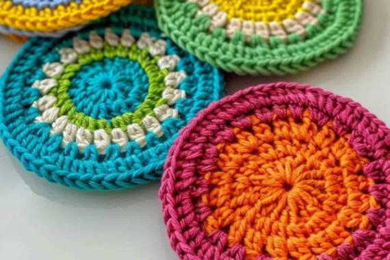 Crocheted coasters in vibrant colors, including orange, pink, blue, green, and yellow, arranged overlapping on a light surface. Bright and playful.