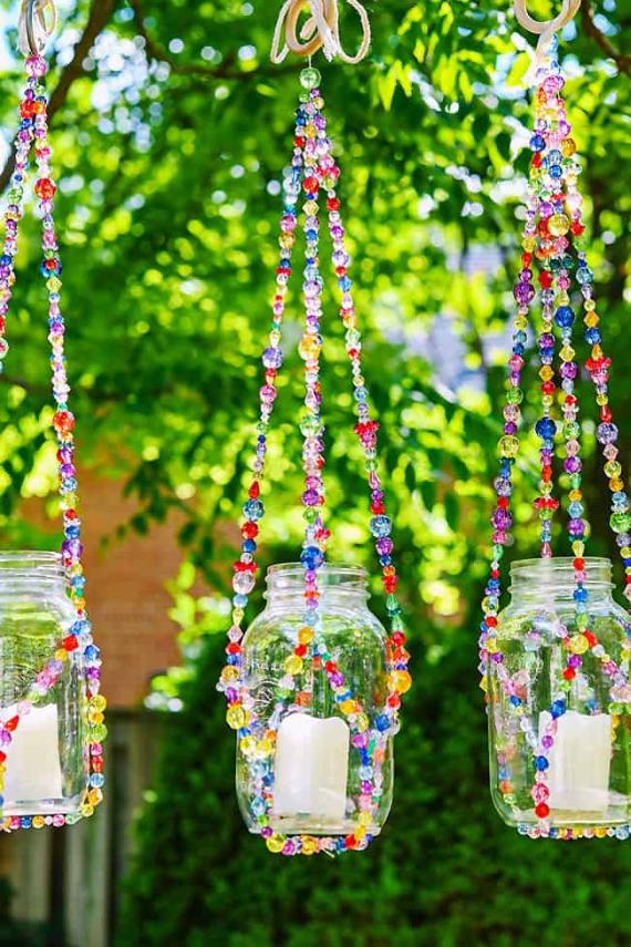 Three glass jars with candles inside are hanging outdoors. They are suspended by colorful beaded strings, with lush green trees in the background.