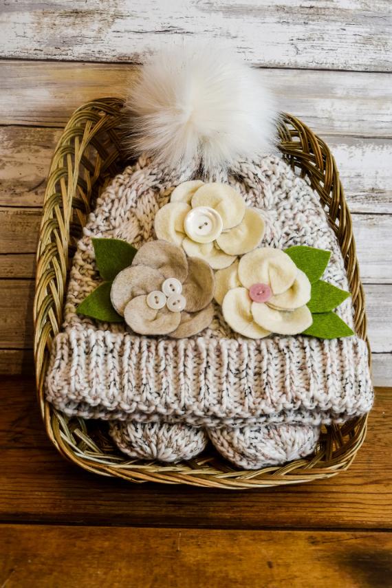 Knitted hat with pom-pom and felt flower appliqués in a wicker basket, set against a rustic wooden backdrop. Warm, cozy, and inviting.