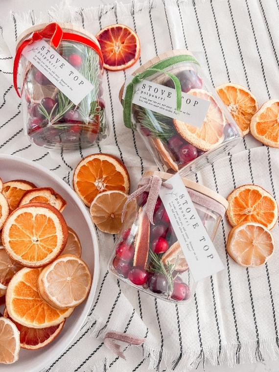 Three festive jars with red, green, and beige ribbons contain dried oranges, cranberries, and rosemary. Nearby, dried citrus slices are scattered on a striped cloth.