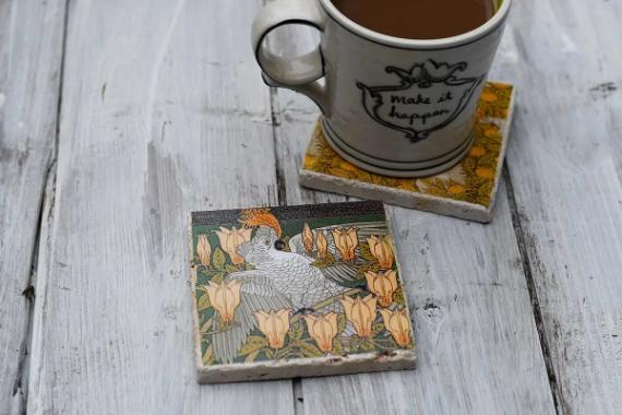 A ceramic mug with coffee on a sunflower coaster. Nearby, a decorative tile features a cockatoo amidst orange flowers on a rustic wooden surface.