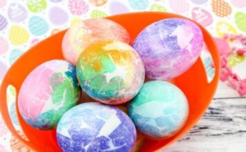 Easter Egg Decorating Ideas – Easter Egg Crafts A vibrant orange bowl holds six colorful tie-dye Easter eggs/ tissue Paper Mosaic Eggs in pastel shades of blue, pink, green, and purple. The background features a colorful, patterned design.