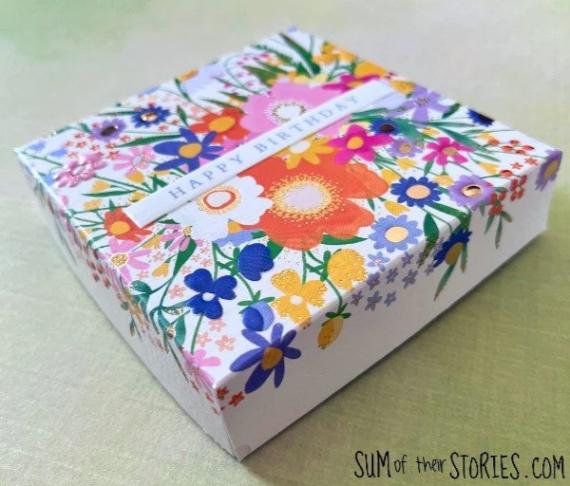 A colorful gift box with "Happy Birthday" text. The box features a vibrant floral pattern with flowers in pink, orange, blue, and yellow hues.