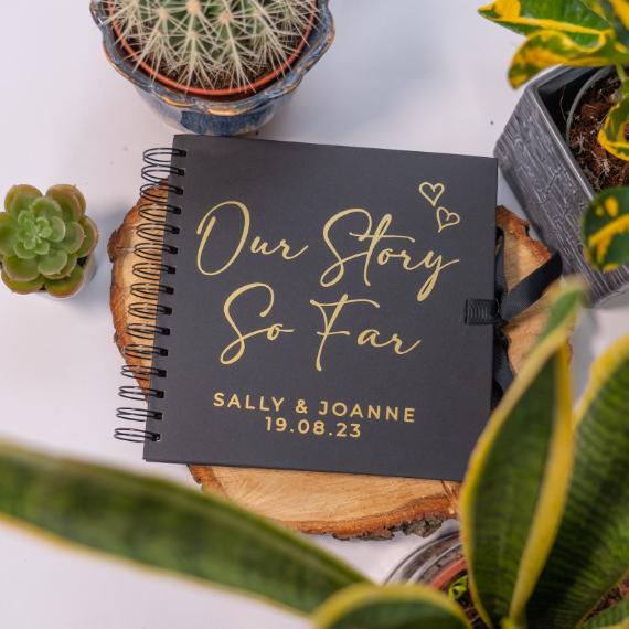 A spiral-bound scrapbook titled "Our Story So Far" with hearts, placed on a wooden slice. Surrounded by potted succulents and plants.