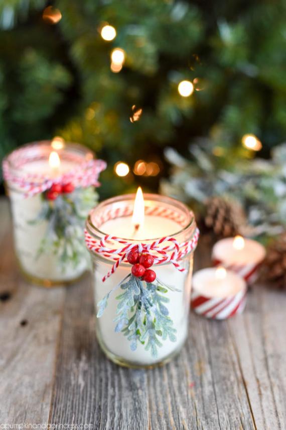 Lit candles in glass jars decorated with red and white twine and pine sprigs sit on a wooden table. A festive, warm atmosphere is created by blurred lights and pinecones.