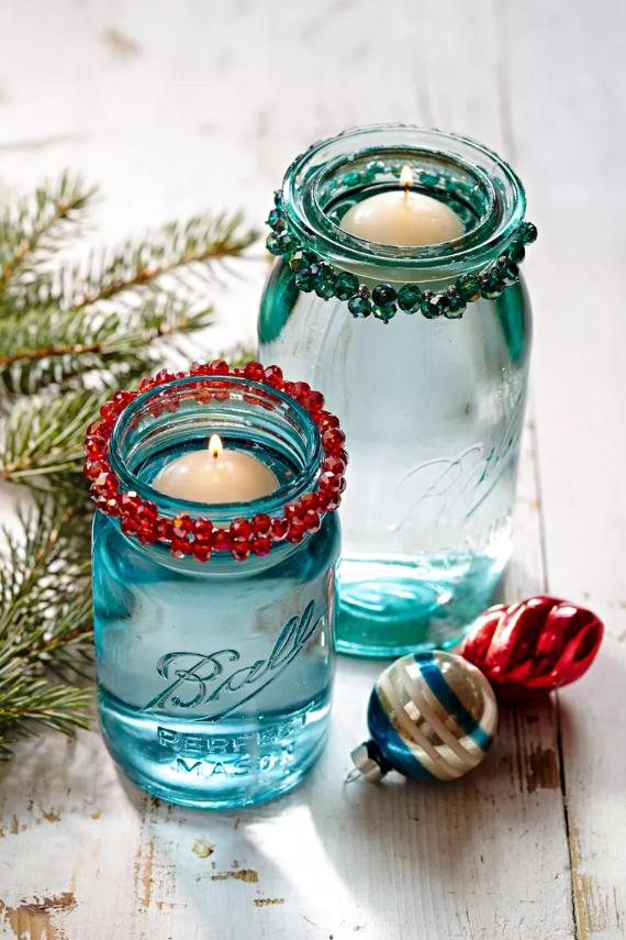 Two teal mason jars with candles, adorned with red and green beaded rims, evoke a festive vibe. Nearby are pine sprigs and colorful ornaments.