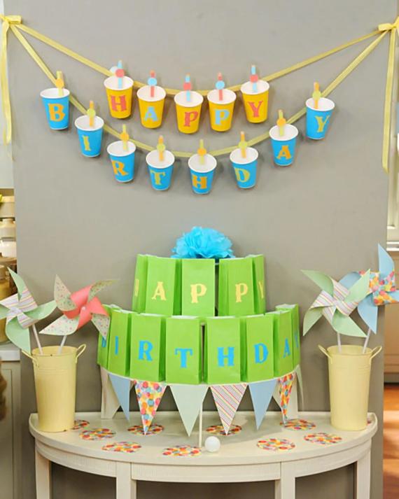 Festive birthday setup with colorful paper cups spelling "Happy Birthday," a green paper cake with a blue pom-pom topper, and decorative pinwheels.