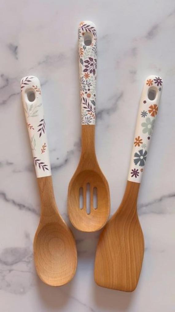 Three wooden kitchen utensils with white floral-patterned handles. The set includes a spoon, a slotted spoon, and a spatula on a marble surface.