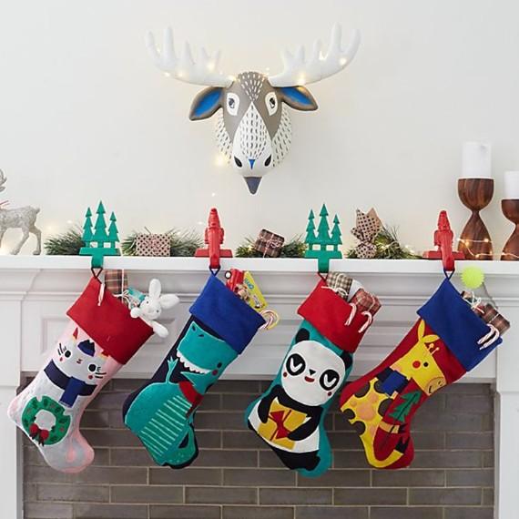 Four colorful Christmas stockings hang from a decorated mantel with lights, gifts, and festive hooks. A whimsical moose head adorns the wall above.
