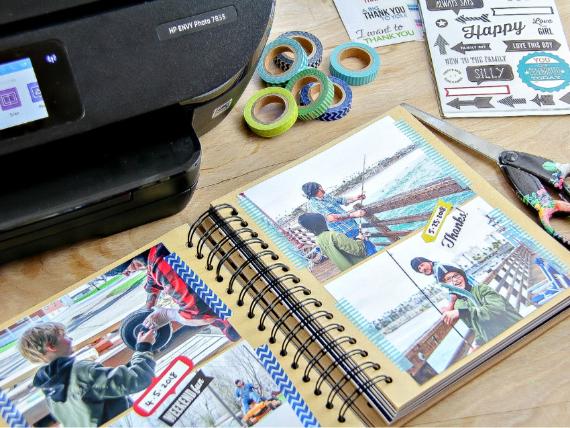 A scrapbook page on a wooden table features fishing photos. Nearby are decorative tapes, scissors, stickers, and a printer, conveying creativity and nostalgia.