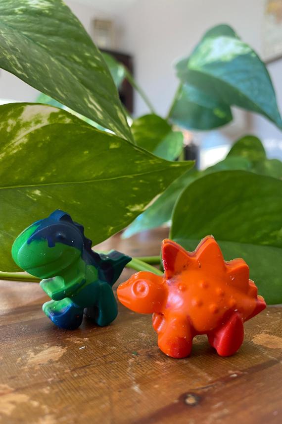 Two colorful toy dinosaurs, one green and blue, the other orange, sit on a wooden surface. Lush green plant leaves create a lively backdrop.