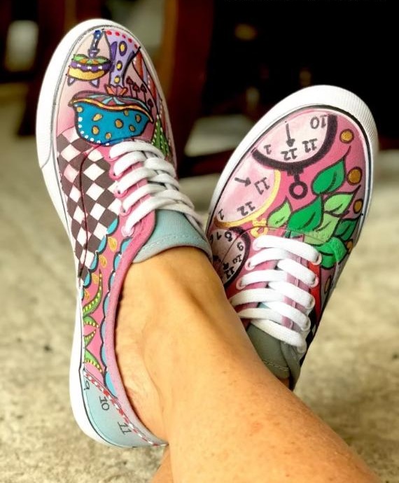 Colorful hand-painted shoes on a person's feet. The designs feature whimsical patterns, clocks, and vibrant shapes, conveying a playful, artistic vibe.