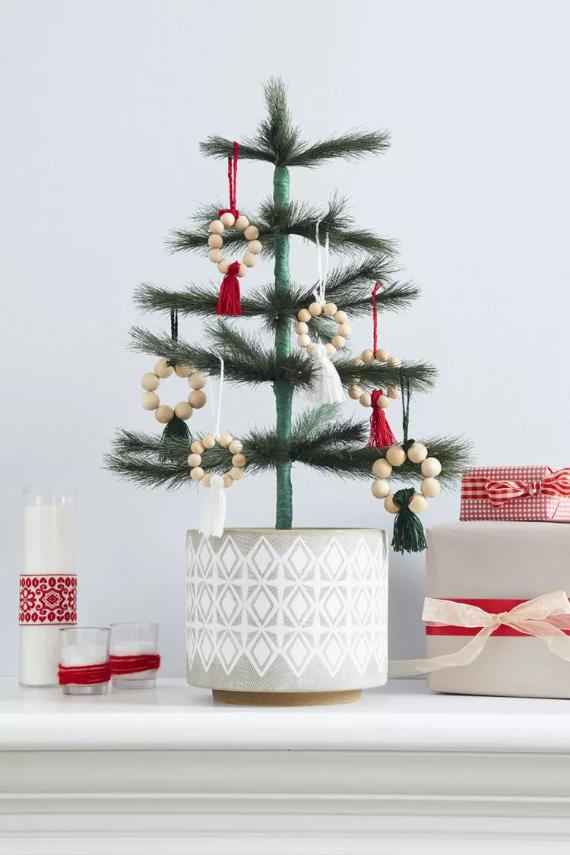 Small Christmas tree in a patterned pot on a mantel, adorned with bead and tassel ornaments. Nearby are white candles and wrapped gifts. Cozy and festive.