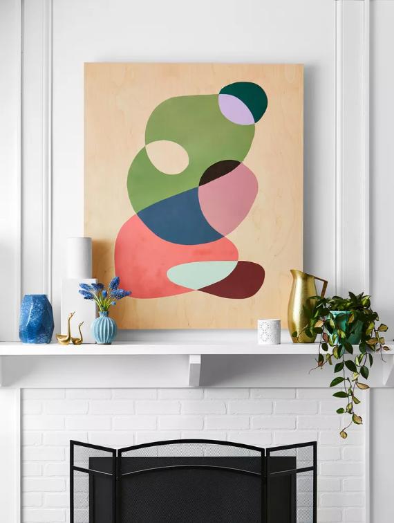 Abstract art with layered geometric shapes, predominantly green and pink, on a wooden panel above a white mantle. Decor includes a blue vase with flowers, a gold watering can, and a potted plant.