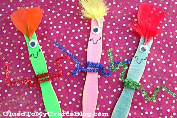 Brightly colored craft sticks with googly eyes and feather hair stand against a pink polka dot background. Each has a pipe cleaner wrapped around. Playful and creative.