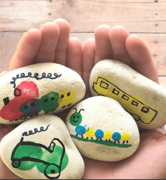 Hands holding four white stones, each painted with colorful vehicles. Designs include a train, bus, car, and caterpillar, creating a playful and creative mood.